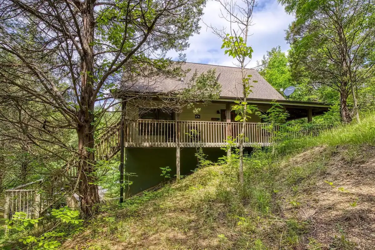 The image showcases the inviting exterior of a charming cabin nestled among lush trees, with a spacious deck perfect for outdoor relaxation. The serene natural setting hints at opportunities for enjoying peaceful sunsets, stargazing, and hosting family gatherings amidst the beauty of the Smoky Mountains.