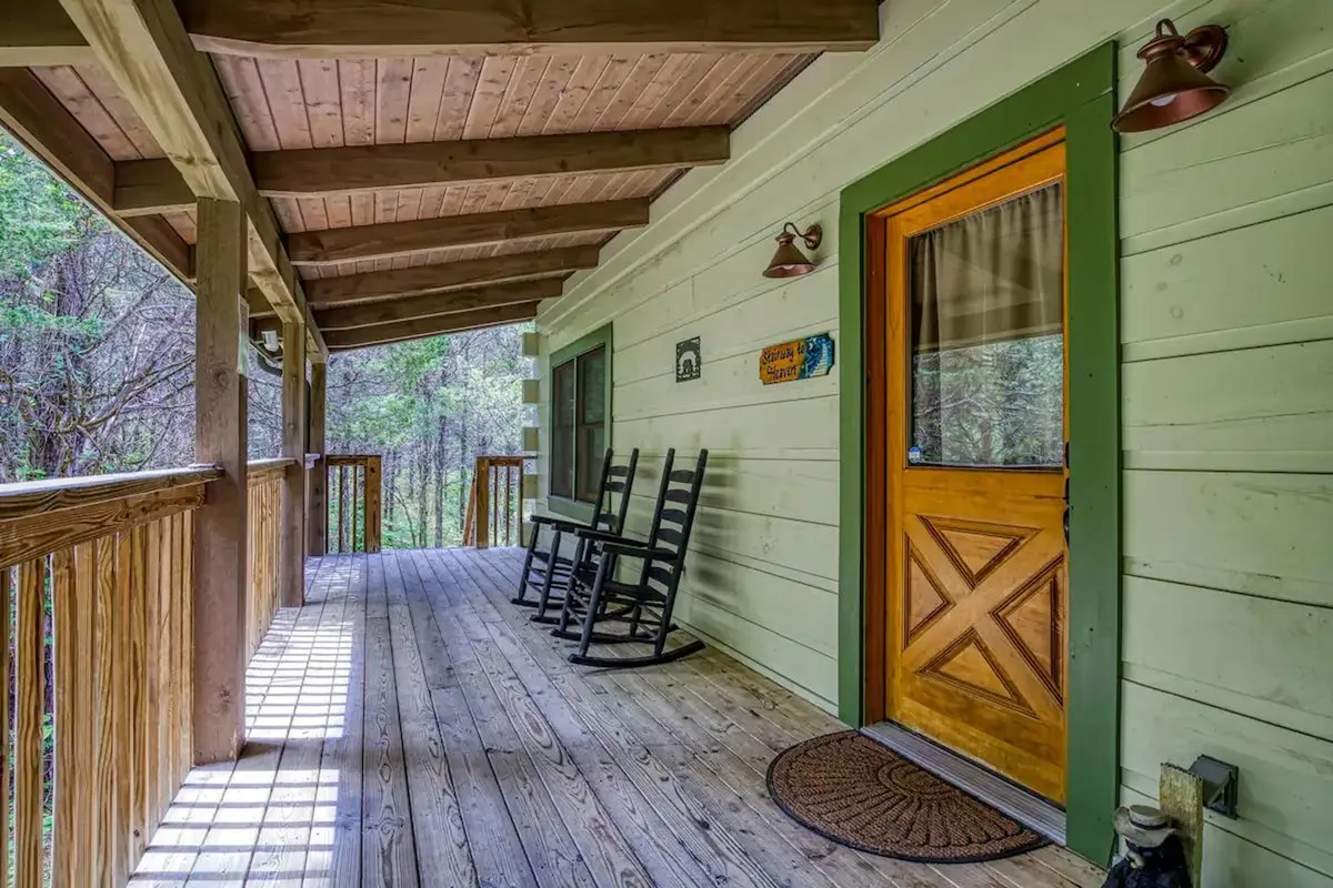 This image features the front porch of the cabin, complete with inviting rocking chairs and a sturdy wooden front door with green trim, all set against a backdrop of lush greenery. The porch looks out over the surrounding woods, offering a tranquil space to enjoy the sights and sounds of nature.