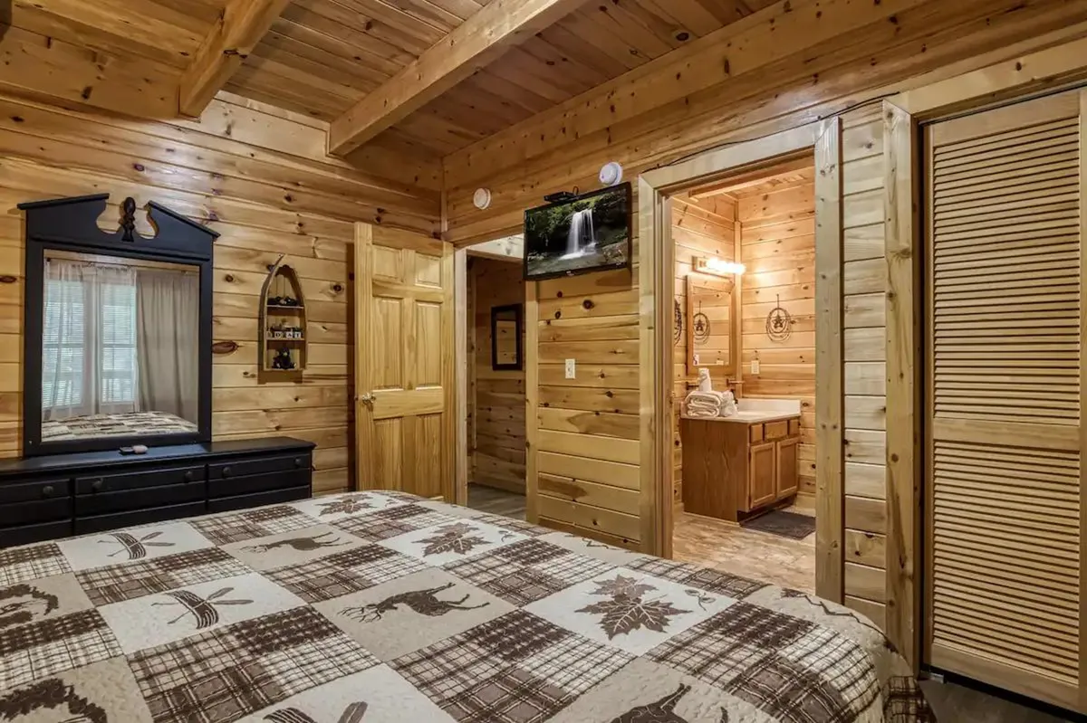 This bedroom image features a wooden bed frame with plaid patterned bedding, a mirror with decorative elements, a wooden dresser, and a doorway that leads to the bathroom. The blend of bedroom and bathroom elements visually supports the convenience of having private bathrooms adjacent to sleeping quarters.
