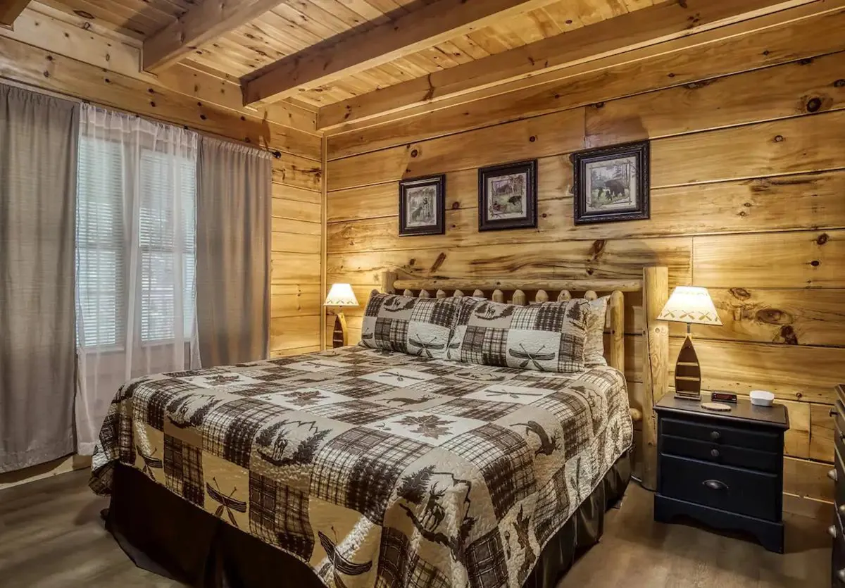 This image showcases a tranquil bedroom with a wooden bed frame, a plaid bedspread, framed artwork above the bed, and nightstands with lamps. The soft lighting and warm wooden paneling create a relaxing, inviting atmosphere—perfect for highlighting the 'plush bedding' and 'tranquil decor' mentioned in the section.