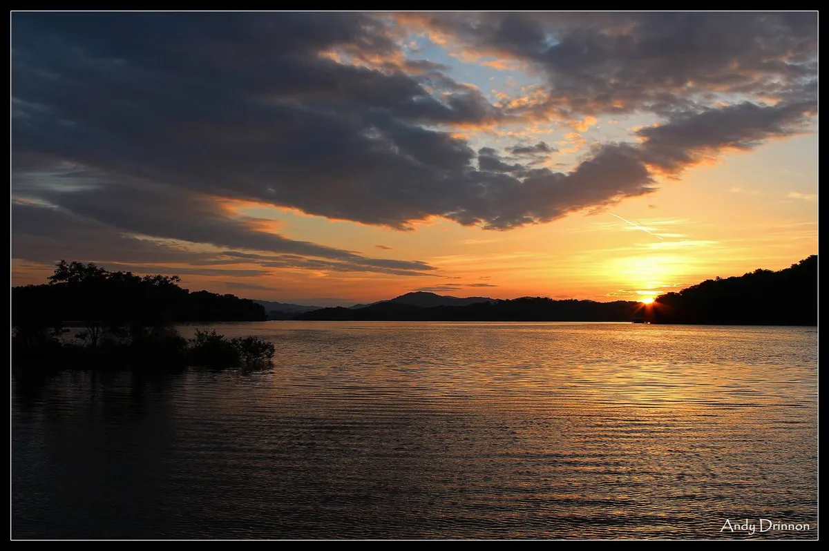 A sweeping view of a tranquil lake at sunset, with calm water reflecting the vivid colors of the sky and distant mountains providing a dramatic backdrop. Gentle cloud formations add to the picturesque serenity. The shoreline hints at peaceful solitude and the potential for romantic moments by the water.
