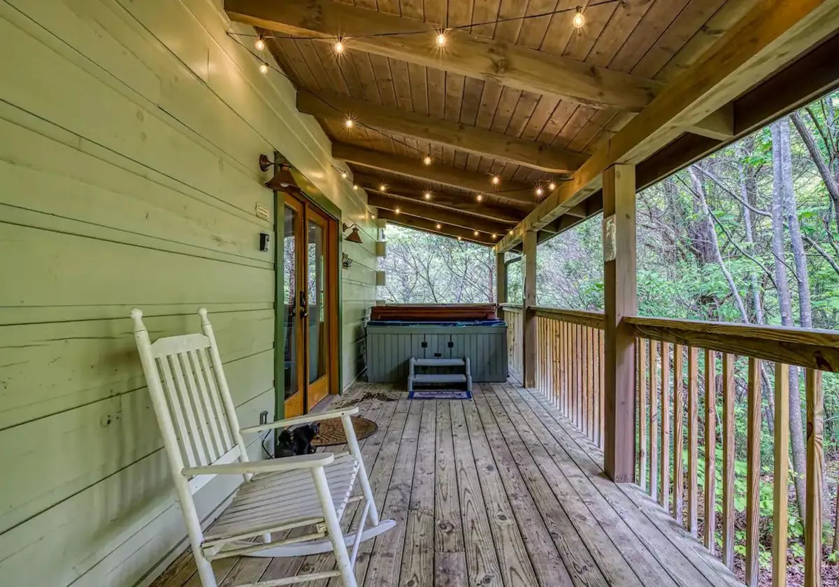 A cozy porch featuring a soothing hot tub tucked into the corner, a classic wooden rocking chair, string lights overhead, and a sturdy wooden railing framing serene views of surrounding greenery. The space is designed for relaxation and gathering, embracing both comfort and natural beauty.
