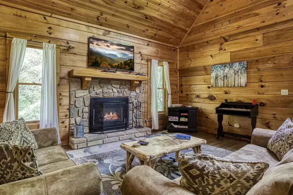 A warmly lit living room with a crackling fireplace as its centerpiece, plush sofas arranged around a coffee table, a large window letting in natural light, and a TV for entertainment. The scene is cozy and inviting, perfect for relaxation, socializing, and board games.