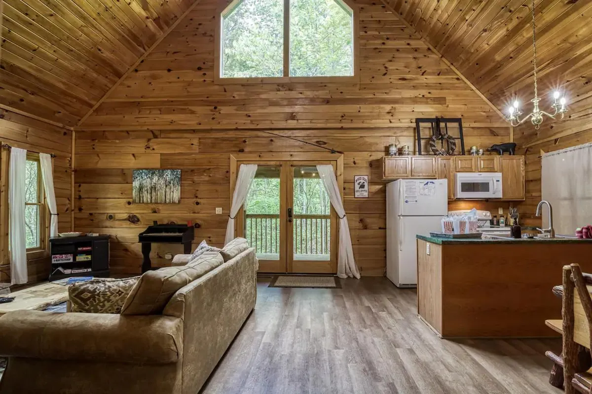 This image presents a wider view of the open-plan living area, where the kitchen area is visible alongside a cozy sofa, large windows, and exposed wooden beams. The open space is bathed in natural light, emphasizing connectivity between cooking and living spaces.
