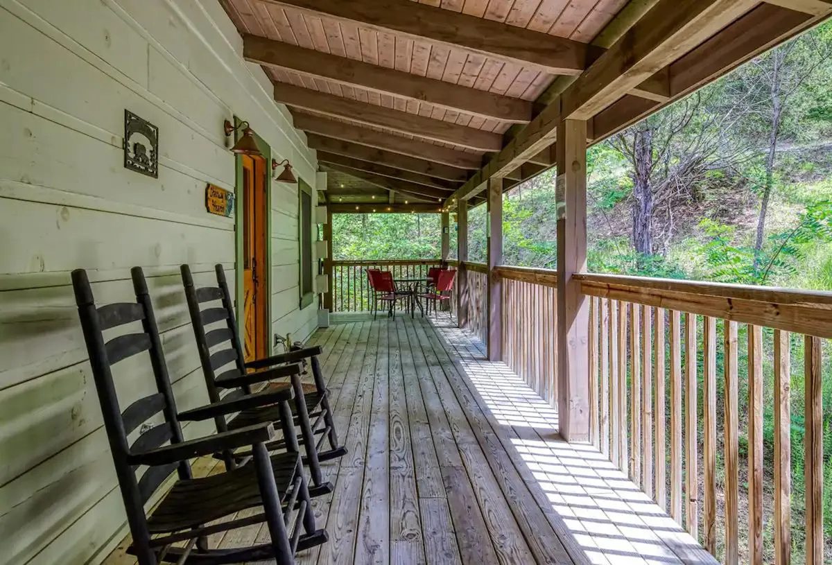 A cozy porch scene with rocking chairs and red chairs arranged along a wooden railing, surrounded by lush greenery. The space suggests a tranquil spot for morning coffee, reading, or enjoying sunset views, directly connecting cabin life to the outdoors.