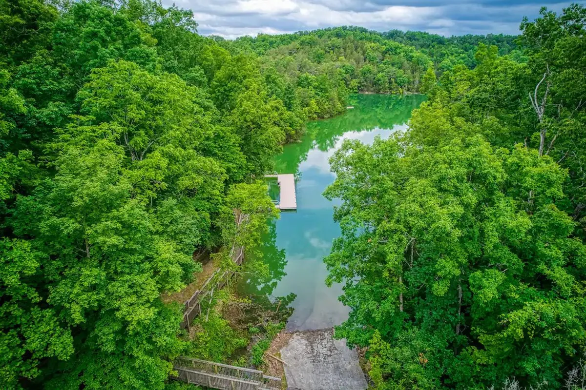 A serene exterior shot of calm water bordered by lush green trees and a wooden dock along the riverbank. The setting invites boating, fishing, picnicking, and nature walks, with the peaceful water reflecting the scenery.