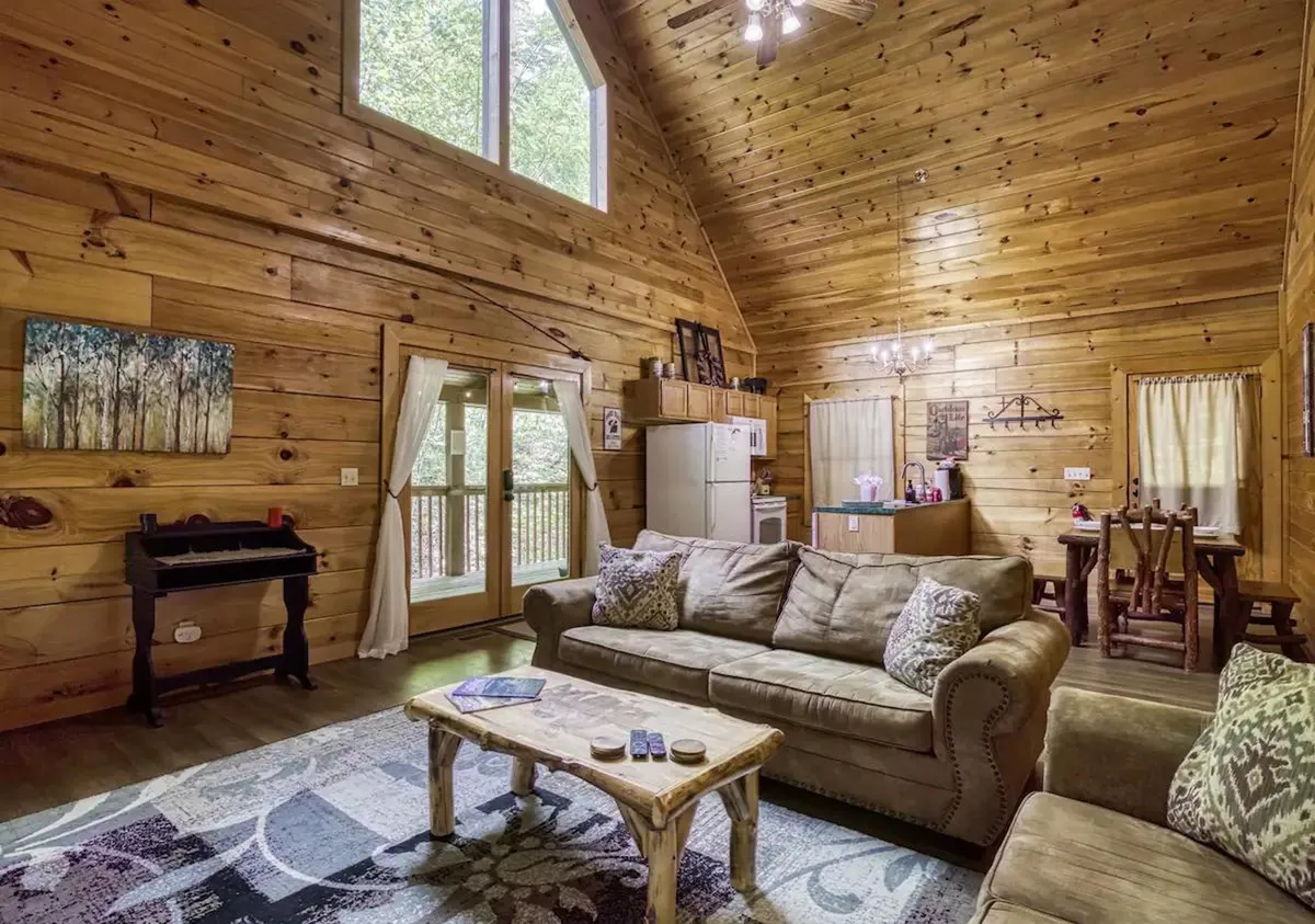 A spacious, open-plan living and dining area with a comfortable sofa, a coffee table, large windows with scenic views, a visible dining space, and a piano in the background. The room is filled with natural light and rustic charm, inviting both relaxation and group gatherings.