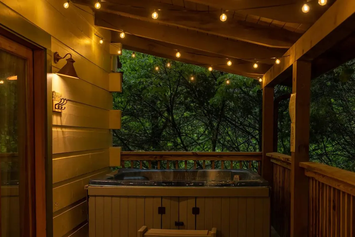 A cozy outdoor setting featuring a hot tub surrounded by string lights and a wooden railing, perfect for relaxing evenings. The ambient lighting and natural surroundings create an inviting atmosphere for socializing under the stars.